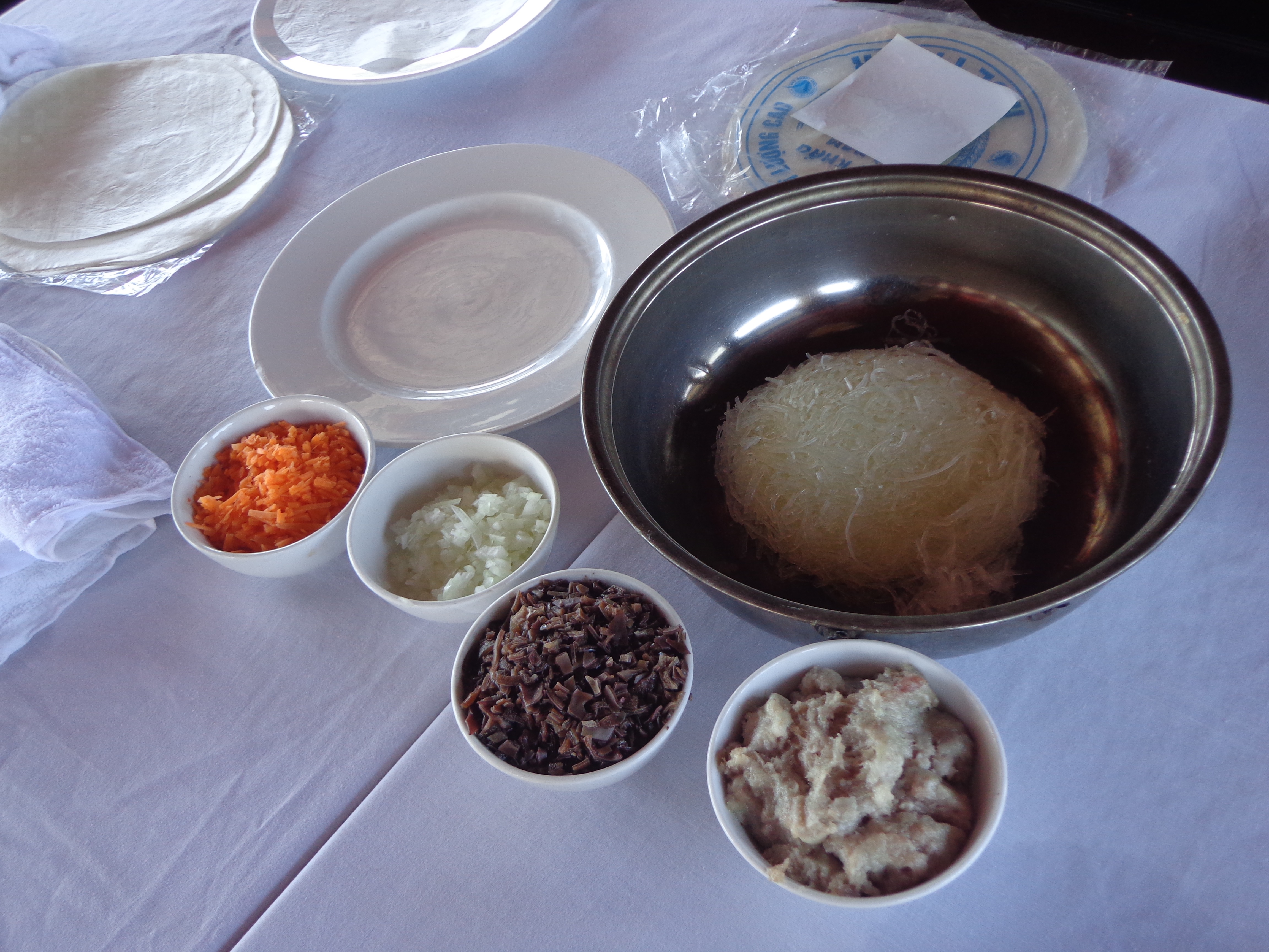 The ingredients to make spring roll. Rice paper on the plate, glass noodles in the steel bowl, carrots, onions, mushroom and cooked pork in smaller bowls.